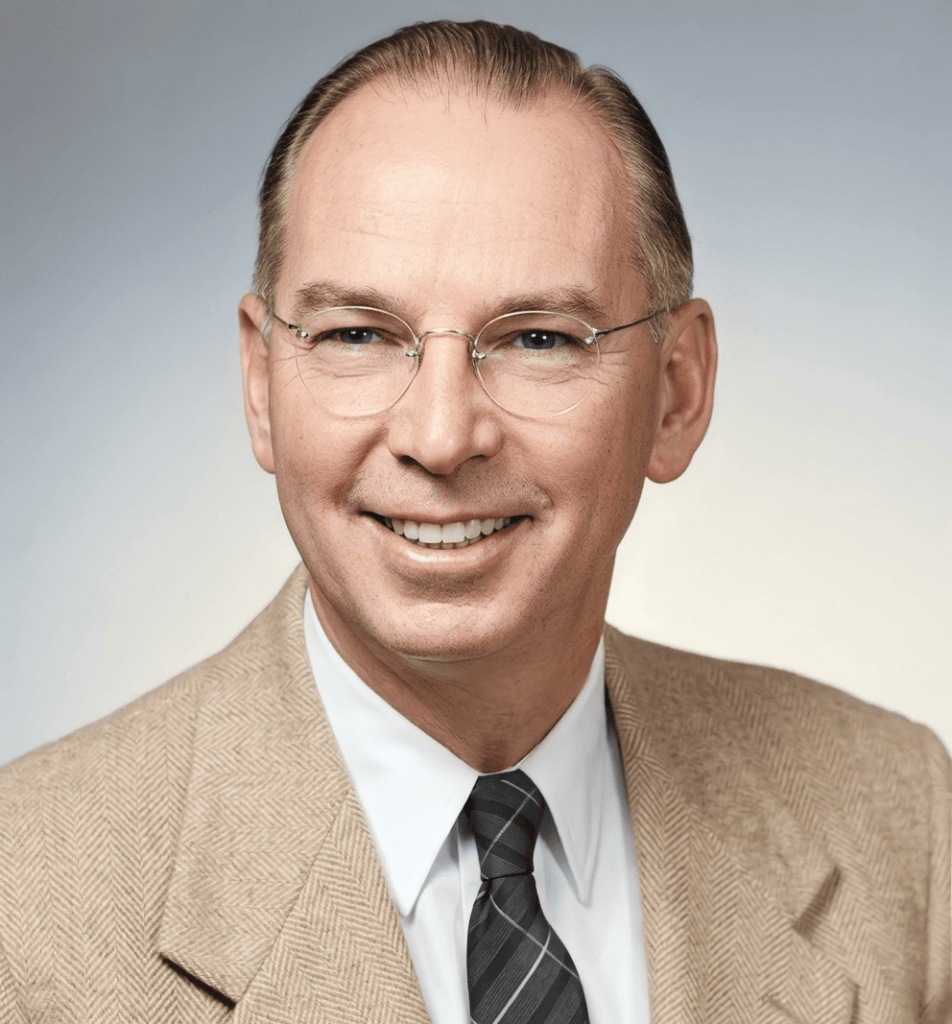 A smiling man wearing glasses, a light-colored suit jacket, and a striped tie, posed against a soft gradient background.