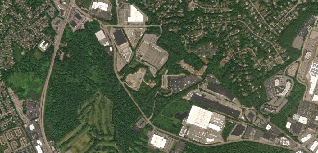 Aerial view of a suburban area featuring a mix of residential neighborhoods, commercial developments, and patches of green space, with roads and parking lots visible throughout.