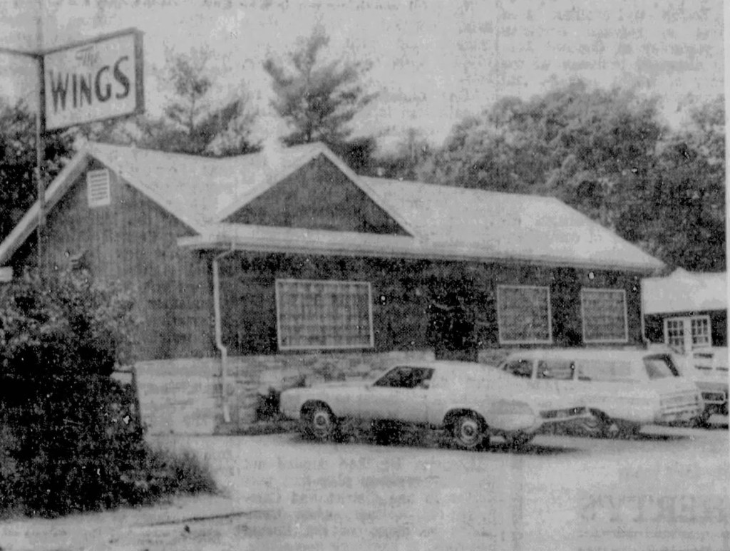 A vintage black and white photograph of a building with a sign reading 'The Wings.' Several cars are parked in front, with trees in the background.