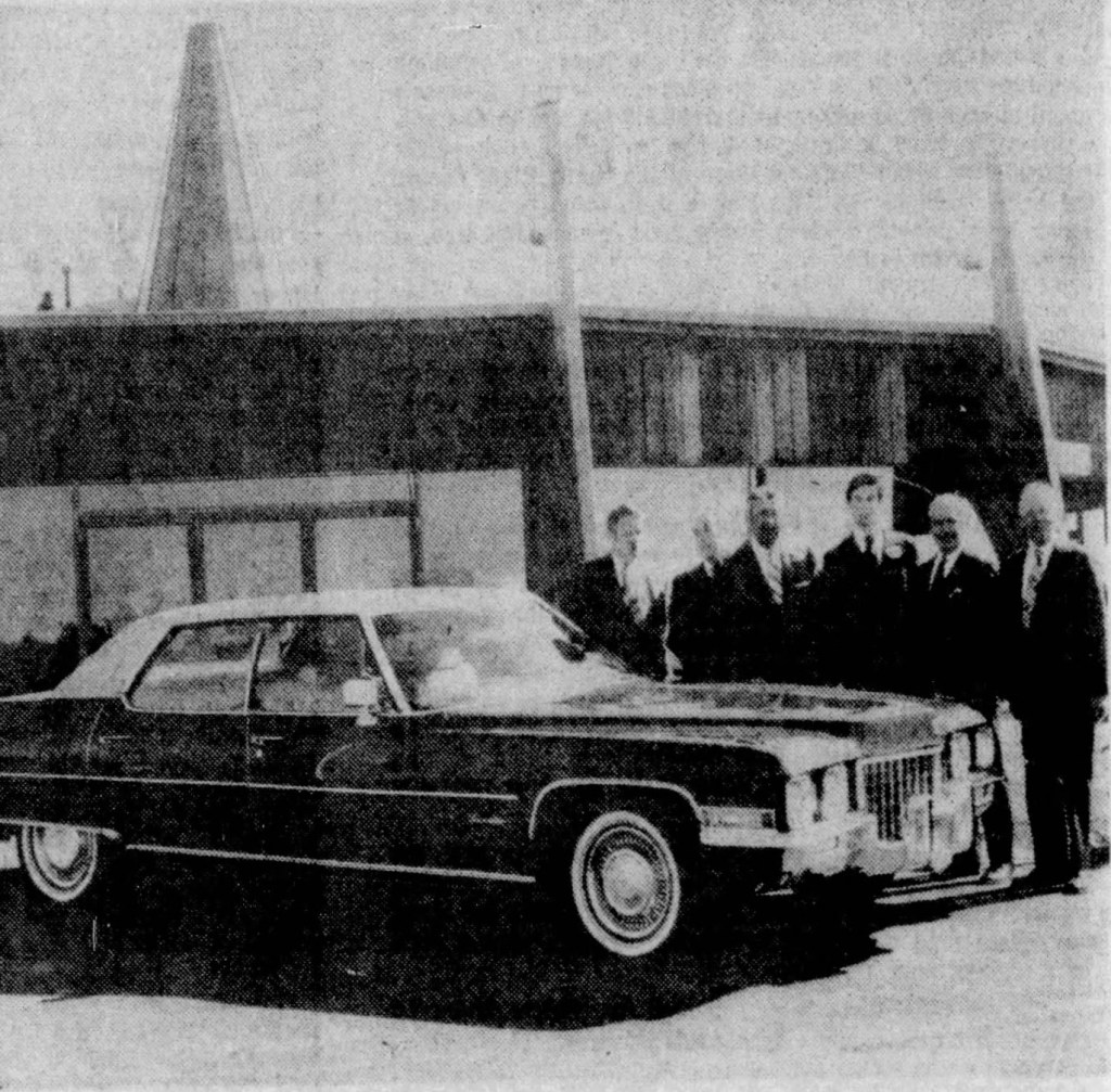 Group of six men in suits standing beside a vintage black Cadillac car in front of a building.