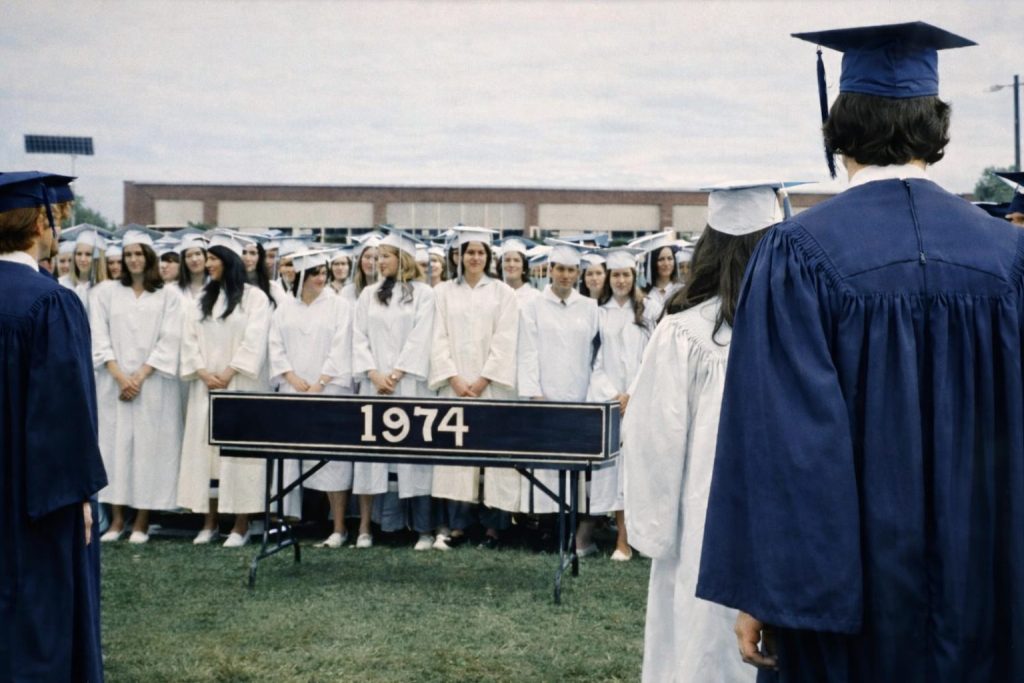 A graduation ceremony in 1974, featuring a diverse group of students in white and blue gowns, standing in formation outdoors, with a sign displaying '1974' in front.