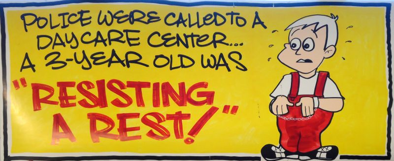 A humorous sign depicting a cartoon boy in red overalls looking concerned, with text that reads 'Police were called to a daycare center... A 3-year-old was "resisting a rest!"' against a bright yellow background.