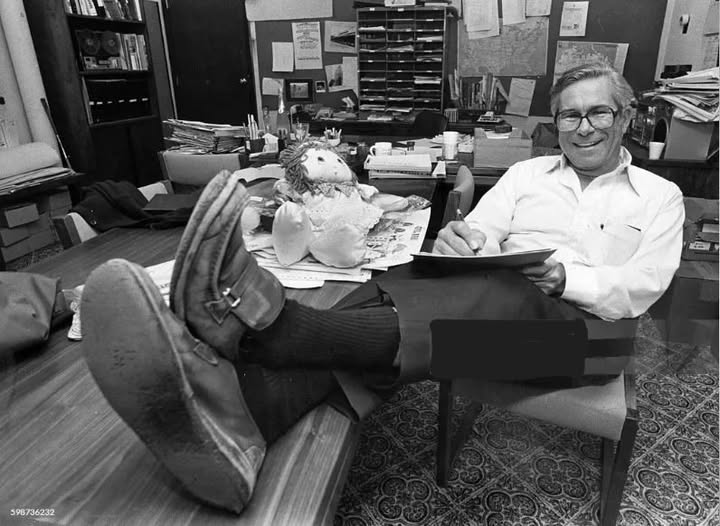 A smiling man sitting at a cluttered desk with his feet resting on the table, wearing a white shirt and dark pants, while holding a notepad. A plush doll is positioned next to him.