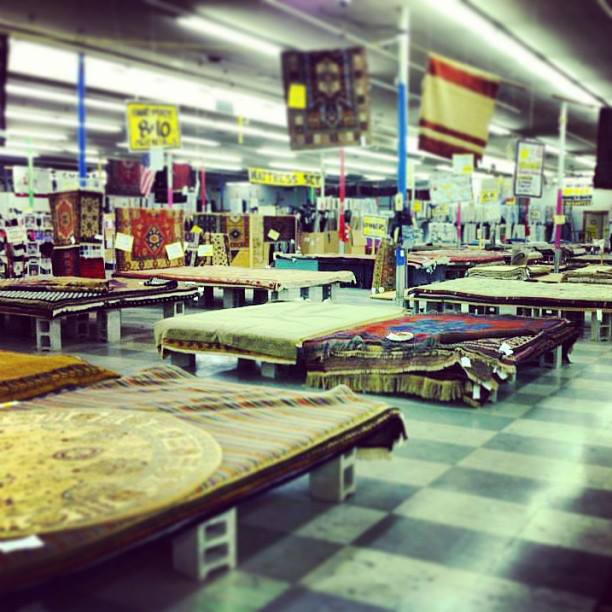 Interior view of a warehouse displaying various rugs and carpets on tables, with colorful signage overhead.