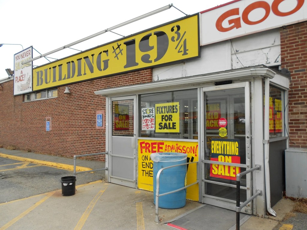 Exterior view of a building with 'BUILDING #19¾' prominently displayed, featuring signs for sales and free admission, alongside a blue barrel and a handicap-accessible ramp.