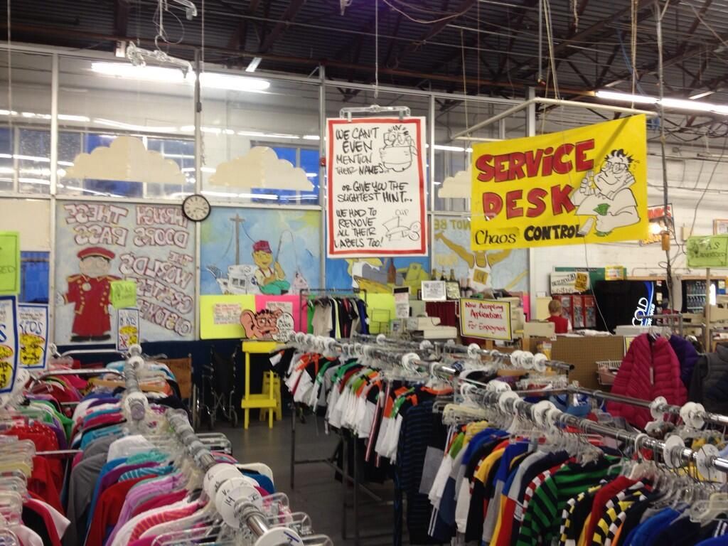 Interior view of a thrift store with colorful clothing racks, signs on the walls, and a service desk labeled 'Service Desk - Chaos Control'.
