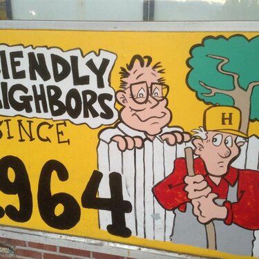 A colorful sign featuring two cartoon characters, one peering over a fence and the other holding a tool, with the text 'Friendly Neighbors Since 1964'.