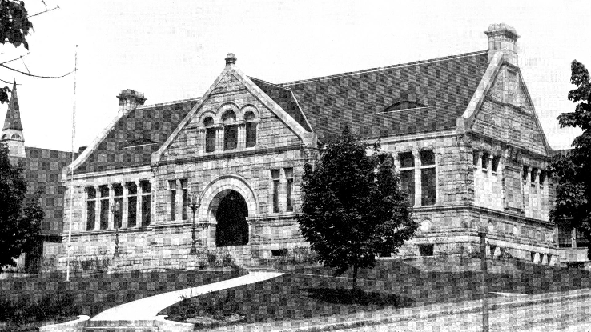 Historic public library building with stone exterior and decorative elements, surrounded by landscaped grounds.