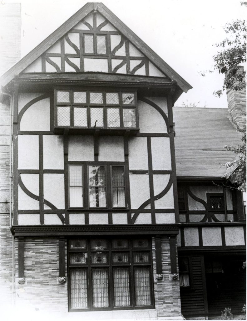 A black and white photograph of a Tudor-style house featuring decorative wooden framing, multiple windows, and a steep roof.
