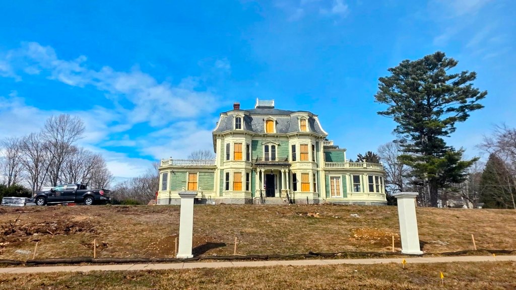 A large, Victorian-style house painted in light green with orange shutters, standing on a grassy hillside under a blue sky. In the foreground, a white stone post is visible, with a truck parked nearby and trees in the background.