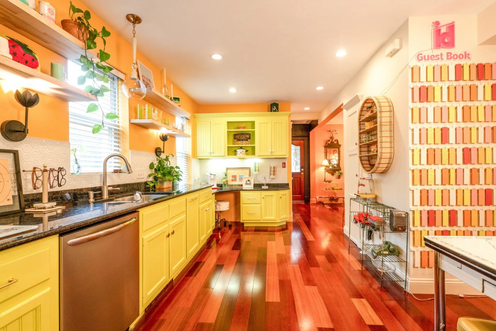 Brightly colored kitchen featuring yellow cabinets, a granite counter, and wooden flooring. Shelves with plants and decorative items are seen along the walls, alongside a unique guest book display.