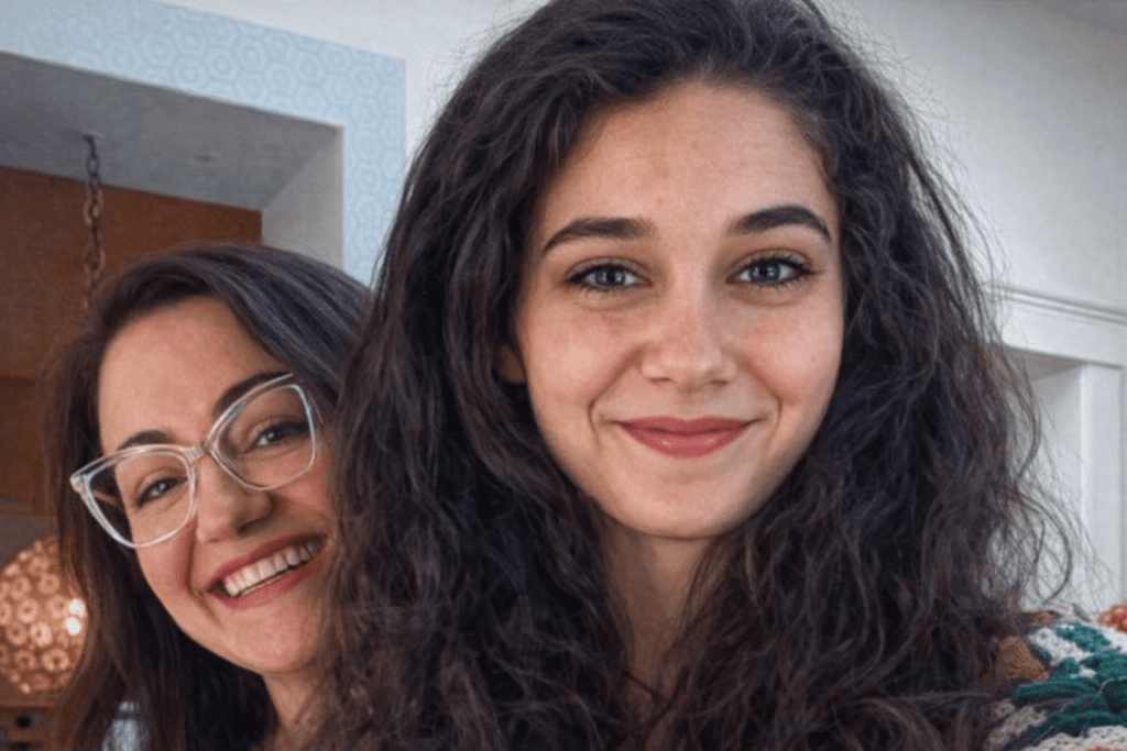 Two women smiling together for a selfie, one with glasses and the other with curly hair, in a well-lit indoor setting.