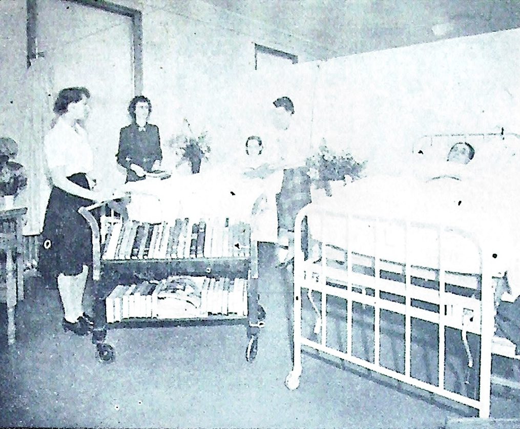 Black and white photograph of a hospital scene with female nurses and a patient in bed. One nurse is pushing a cart filled with books, while another nurse is attending to a patient. A woman stands nearby, and flowers are located in the background.