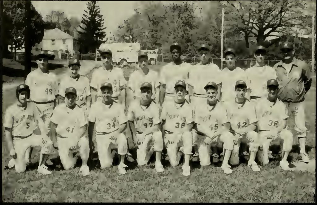 A black and white photo of a baseball team posing on a grassy field, featuring players in uniforms with numbers, standing and sitting in two rows. Trees and a vehicle can be seen in the background.