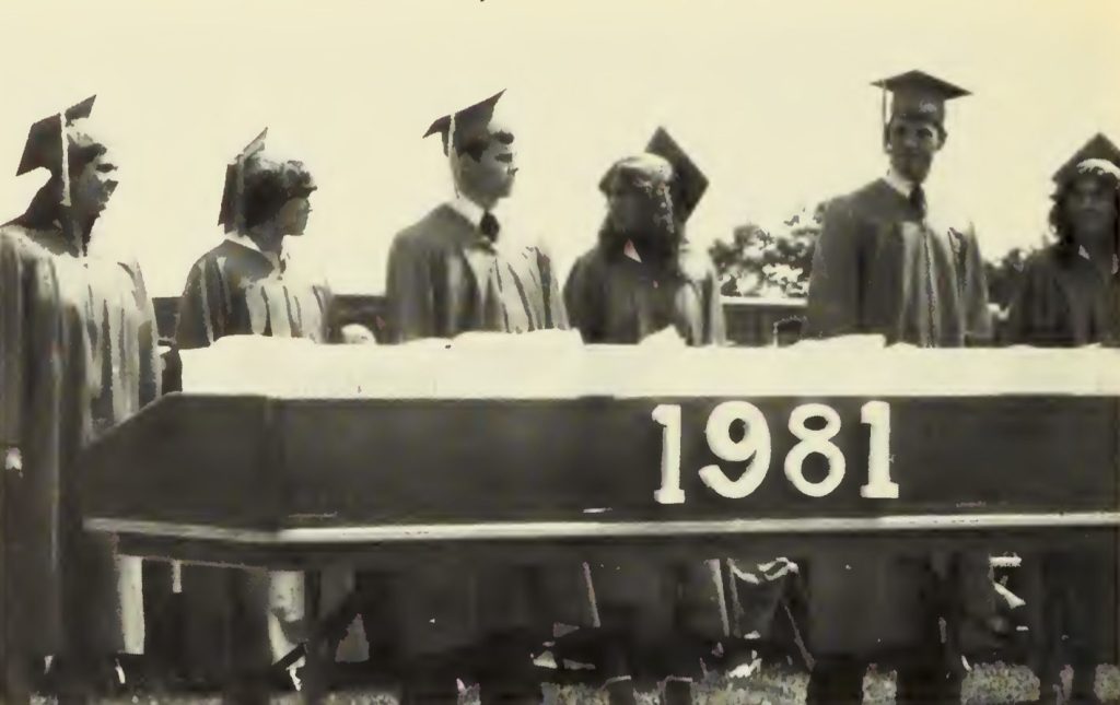 Students in graduation gowns and caps standing in a row, with a sign displaying '1981' in front of them.