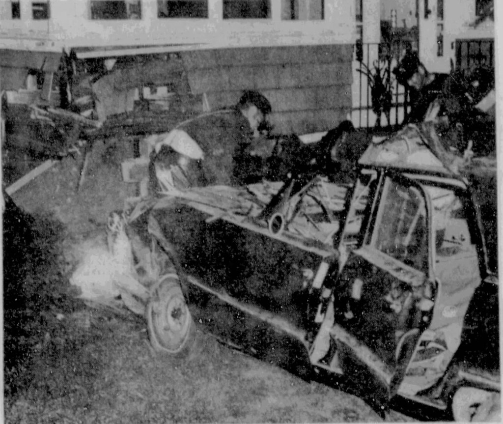 A damaged car is shown partially on a lawn near a house, with individuals examining the wreckage.