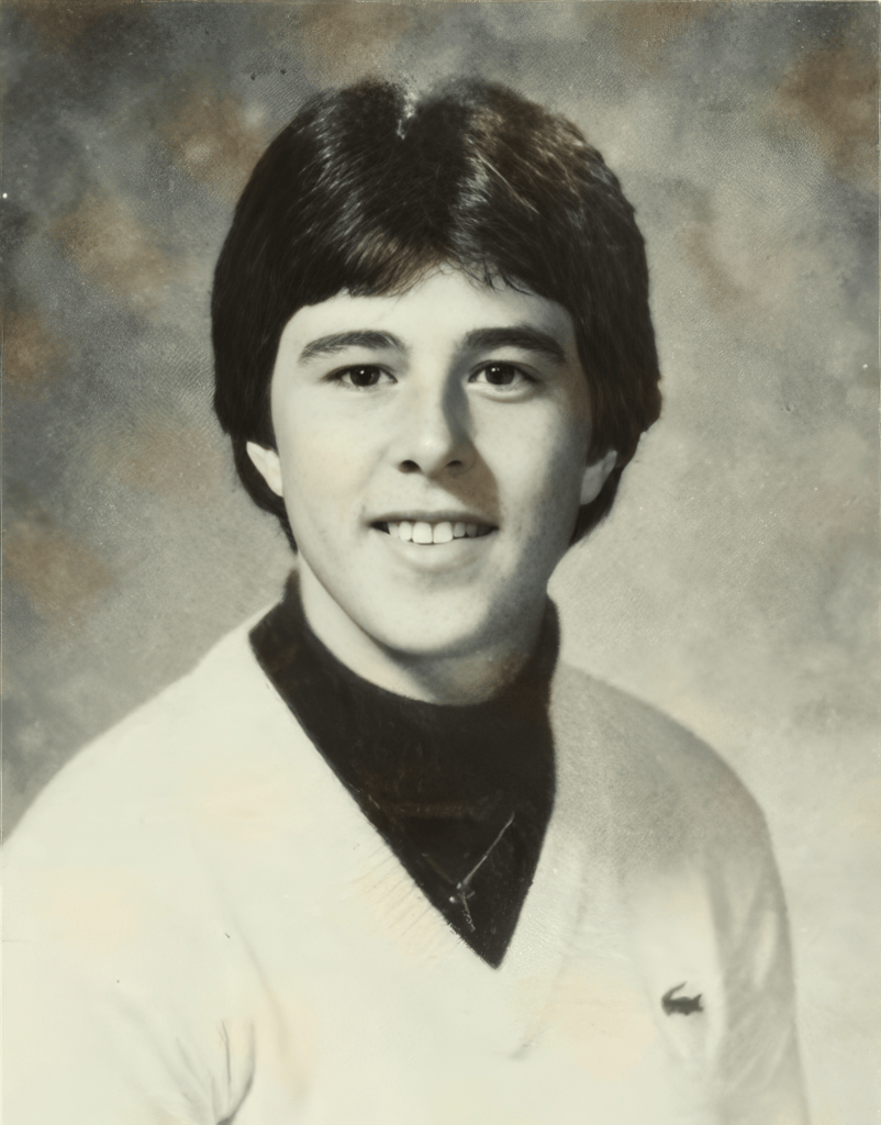 A vintage portrait of a young man with dark, styled hair, smiling at the camera. He is wearing a white sweater over a black turtleneck, with a simple necklace visible. The background is a soft gradient, creating a classic school photo aesthetic.