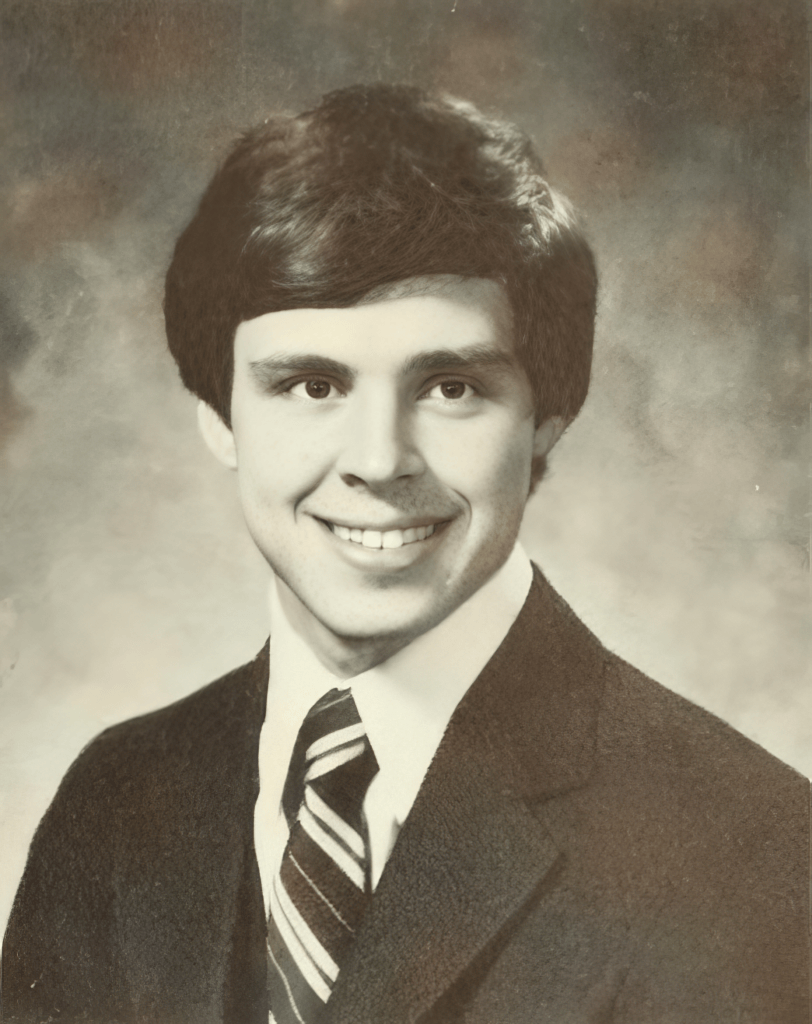 A young man with dark hair and a friendly smile, wearing a suit and tie in a formal portrait setting.
