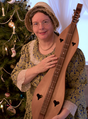 Woman in historical attire holding a mountain dulcimer near a decorated Christmas tree